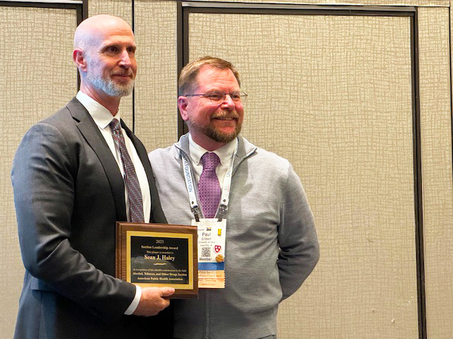 A tall beareded caucasian professor receives a plaque from a shorter, bearded caucasian professor, standing in front of beige conference room walls, which belies the fact that this is in all earnestness an exciting moment for everyone in the room