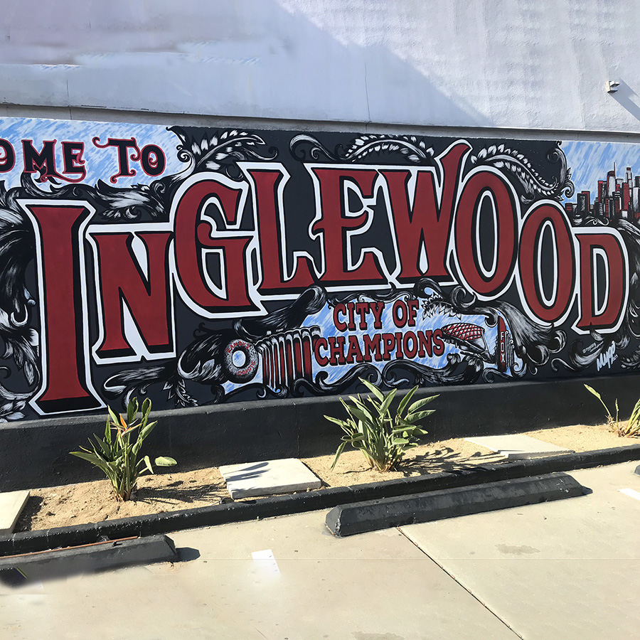 A red, black, and sky blue mural on a gray wall in a parking lot reads "INGLEWOOD" and below it in smaller letters, "CITY OF CHAMPIONS". At the foot of the wall, some succulents grow from tan dirt.