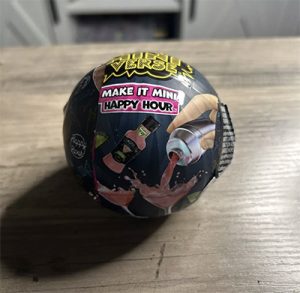 A black plastic-wrapped ball about the size of a baseball sits on a wood table. The black plastic has a pink label that reads "make it mini HAPPY HOUR" on it.