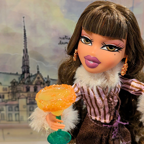 A doll with big hazel eyes, long brown hair with sharp bangs, and a striped tunic with fur sleeves (?) stands in front of a drawing of the Isle de Cite in Paris at sunset, holding a MGA Miniverse "Happy Hour" mango margarita which we had to glue to her hand because it's heavy, but which nonetheless is clearly made at her scale.