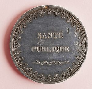 Against a pink-red background, a tarnished bronze medal with an ornate filigreed border reads "SANTÉ PUBLIQUE" 