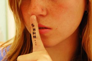 A white preteen girl with holds her index finger up to her lips. Down the finger is written "SHHH...." The image is cropped so you cannot see her eyes.