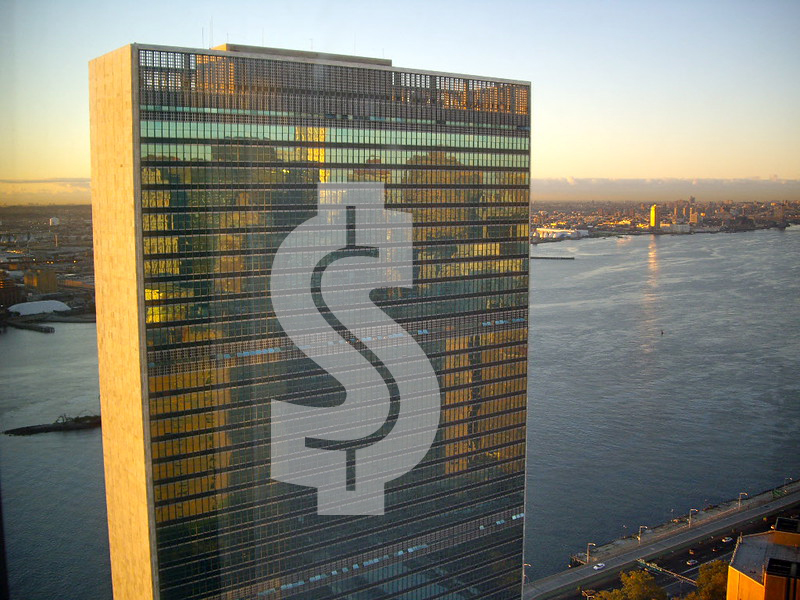 A large square building--the UN tower--stands in front of the blue-grey East River at dawn. The windows are glowing bronze, but there is an enormous dollar sign printed on the side of the building.