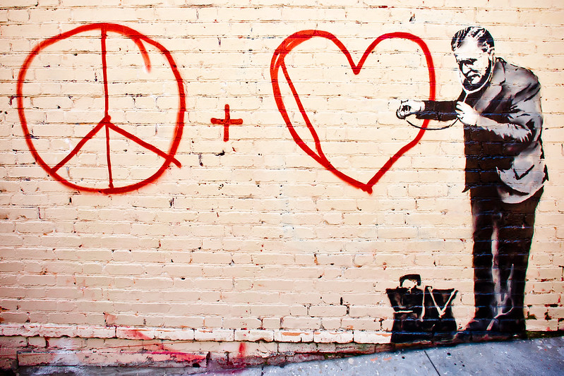 A tan-painted brick wall with a simple red spray-painted peace plus love on it. At the far right, a carefully stenciled old-timey doctor holds a stethoscope up to the heart. The sidewalk is clearly slanted--this is on a steep hill.
