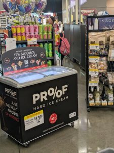 On a gray linoleum super market floor, a big black freezer chest says "PROOF" on it with the two O's arrange like a percentage sign.  A store-placed price sign say "GREAT CHOICE!" Behind it, in the checkout line, you can see the pink backpack of a young girl and the outline of her mom.