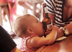 A close-cut photo across a wooden bar top, of a baby seated next to an adult in a striped shirt. The baby has grabbed a long-necked bottle of beer and is pressing the side to its face. 